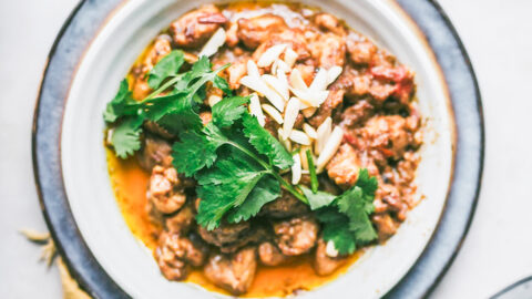 bowl of pakistani korma garnished with parsley and almond and surrounded by whole spices used in cooking