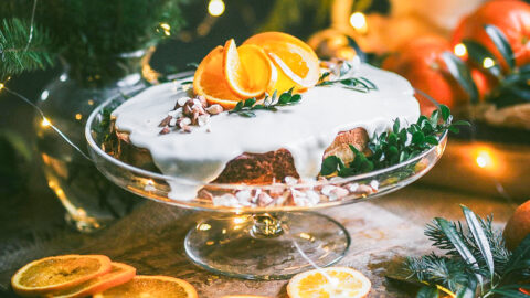 gorgeous almond vasilopita sits on glass cake stand. garnished with decorative orange slices and artful drips of glaze.