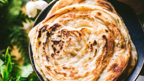 hand holds flaky spiraled roti paratha (aka roti canai in malaysia) above lush green foliage.