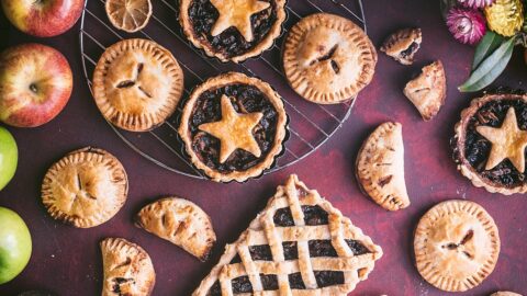 festive overhead shot of christmas mincemeat pies in all shapes and sizes on red background. lattice rectangle tarts, star shaped round tart, handheld pastries. surrounced by apples, dried lemons, flowers, and greenery